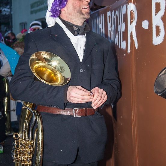 Ein Mann mit lila Haaren und einer lila Perücke spielt eine Tuba bei einer Karnevalsveranstaltung.