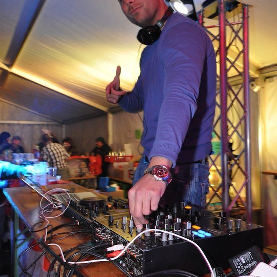 Ein Mann in einem lila Hemd gibt ein Daumen hoch, während er einen DJ-Mixer in einem Zelt bedient, mit anderen Leuten im Hintergrund.