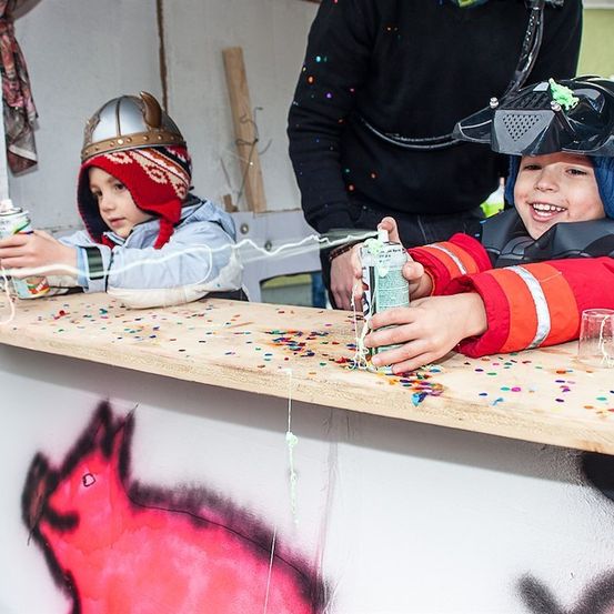 Zwei Kinder mit Helmen, eines mit einem gehörnten Helm, sprühen Farbe auf einen Holztresen mit Konfetti. Ein Mann in Schwarz steht hinter ihnen.