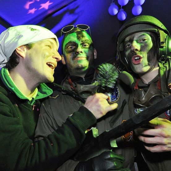 Drei Männer in Militäruniformen mit grüner Gesichtsbemalung und Helmen, einer hält ein großes Objekt, ein anderer eine Gasmaske und ein dritter mit einem weißen Bandana, posieren für ein Foto in einem schwach beleuchteten Raum.