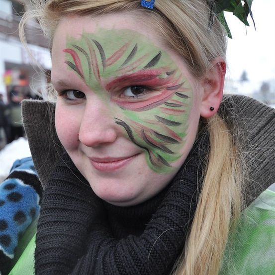 Eine Frau mit langen blonden Haaren und Gesichtsbemalung lächelt in die Kamera, trägt einen schwarzen Schal und eine grüne Jacke.