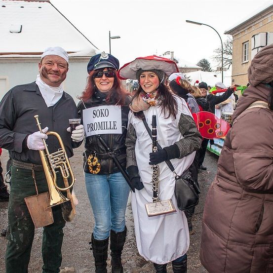 Drei Menschen in Kostümen, einer hält eine Trompete, ein anderer ein Schild mit der Aufschrift 'SOKO PROMILLE', und ein dritter mit einer Klarinette, stehen auf einer verschneiten Straße.