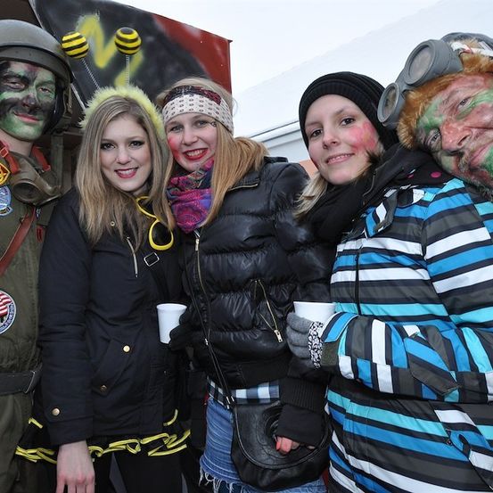 Fünf Menschen stehen zusammen und lächeln für ein Foto. Drei von ihnen sind verkleidet, einer in einer Militäruniform, ein anderer mit einem bienenartigen Outfit und der letzte mit einem gemaltem Zombie-Gesicht. Die anderen beiden sind in Winterkleidung.