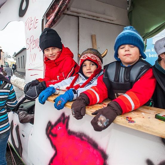 Drei Kinder in Winterkleidung und Wikingerhelmen sitzen in einem dekorierten Fahrzeug, möglicherweise bei einem Festival. Ein Mann mit einem grauen Hut steht hinter ihnen.