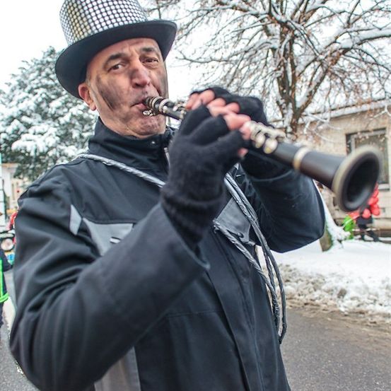 Ein Mann mit einem karierten Hut und schwarzen Handschuhen spielt eine Klarinette in einer verschneiten Außenumgebung.