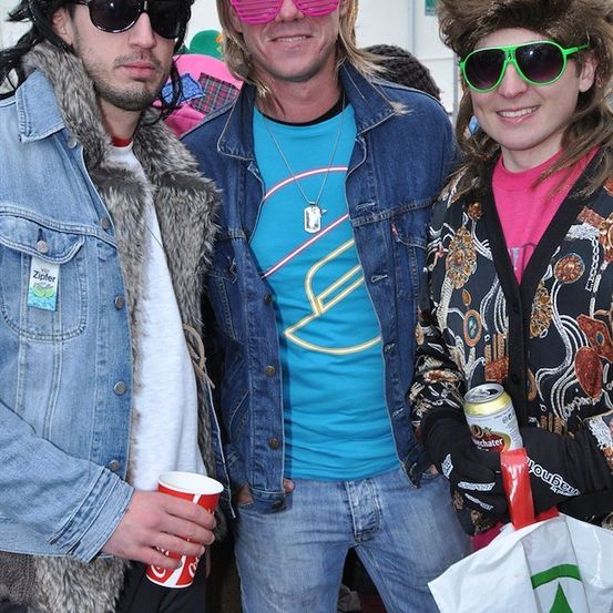 Drei Menschen in 80er-Jahre-Kleidung, einer hält eine Bierdose und einen Plastikbecher, ein anderer eine weiße Tasche mit grünem Logo, der dritte trägt eine Sonnenbrille und eine Halskette.