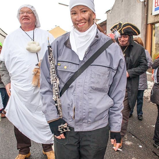 Eine Frau in einer grauen Jacke und schwarzen Hosen steht auf der Straße, hält ein Oboe, mit einem Mann in einem weißen Gewand und einem anderen Mann in einem Piratenhut im Hintergrund.