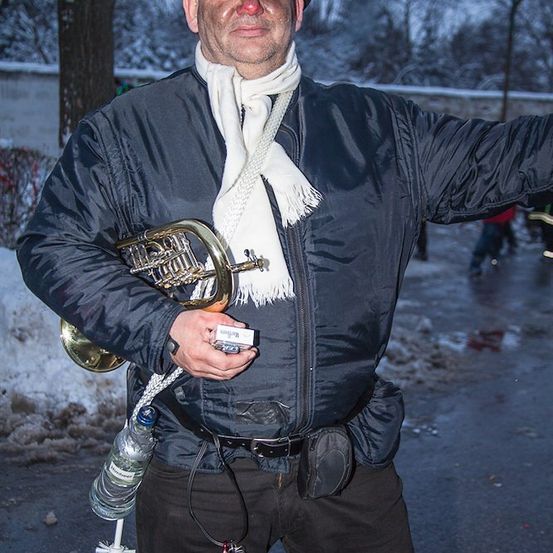 Ein Mann in Winterkleidung, einschließlich Hut, Schal und Jacke, steht draußen im Schnee und hält ein Waldhorn und eine kleine Packung.