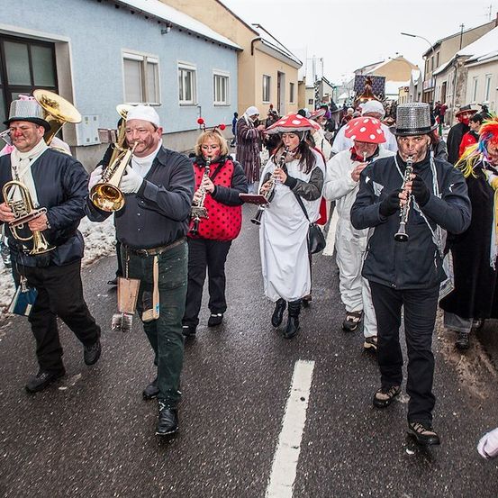 Eine Gruppe von Menschen in Kostümen, die Musikinstrumente spielen, marschiert eine verschneite Straße entlang.