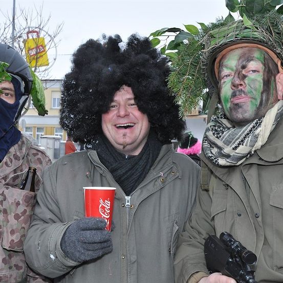 Drei Männer in Militärkleidung, einer mit einem Coca-Cola-Becher, ein anderer mit einem getarnten Helm und Gesichtsbemalung, und der dritte mit einem Helm, der mit Blättern geschmückt ist.