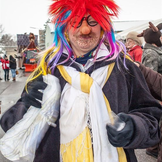 Eine Person mit einer farbenfrohen Perücke und Gesichtsbemalung, gekleidet in einem Kostüm mit einem gelben Schal, hält eine Plastiktüte. Im Hintergrund sind Menschen in Winterkleidung und eine große Struktur zu sehen.