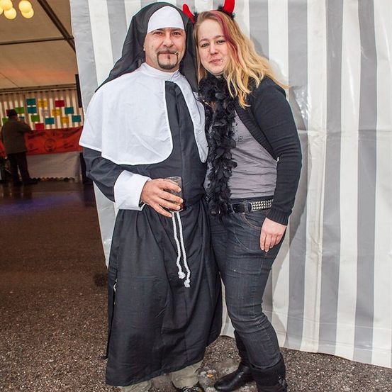 Ein Mann, der als Nonne gekleidet ist, und eine Frau mit roten Haaren und einem schwarzen Federboa stehen zusammen vor einem gestreiften Hintergrund.