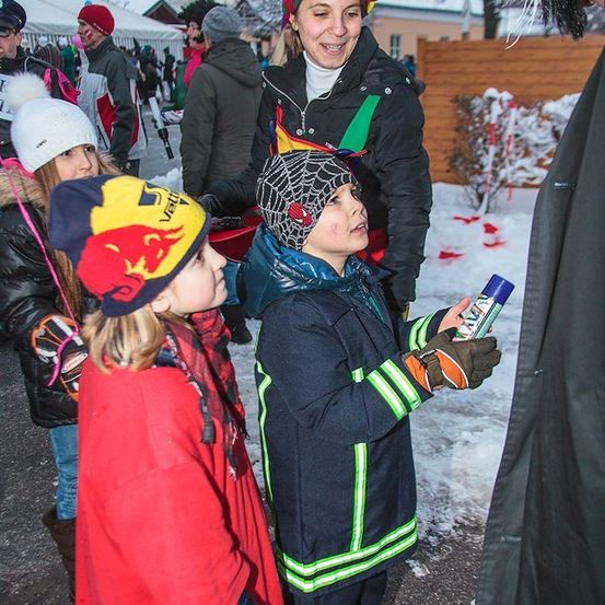 Eine Gruppe von Kindern und Erwachsenen in Winterkleidung, einige mit Mützen, stehen in einem verschneiten Bereich, eine Frau hält eine blaue Flasche und ein Junge in Feuerwehruniform schaut sie an.
