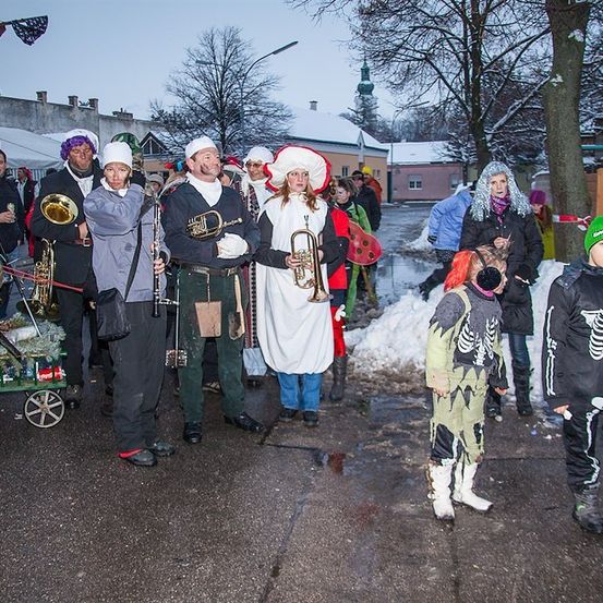 Eine Gruppe von Menschen in verschiedenen Kostümen, darunter ein Skelett, steht auf einer verschneiten Straße. Einige halten Musikinstrumente.