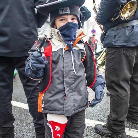 Ein kleiner Junge, der einen Cowboyhut, eine Mütze und Handschuhe trägt, hält eine Spielzeugpistole in der Hand und steht auf einer Straße mit Musikern im Hintergrund.