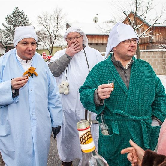 Drei Männer in Roben und Hüten stehen draußen in einer verschneiten Umgebung. Der Mann auf der linken Seite hält ein Kuscheltier, der Mann in der Mitte hat eine Zigarette in der Hand und der Mann auf der rechten Seite hält ein Glas.