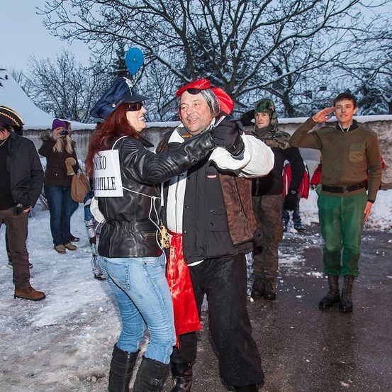 Eine Gruppe von Menschen in Kostümen steht im Schnee, ein Paar tanzt und andere schauen zu. Bäume und ein Gebäude sind im Hintergrund.