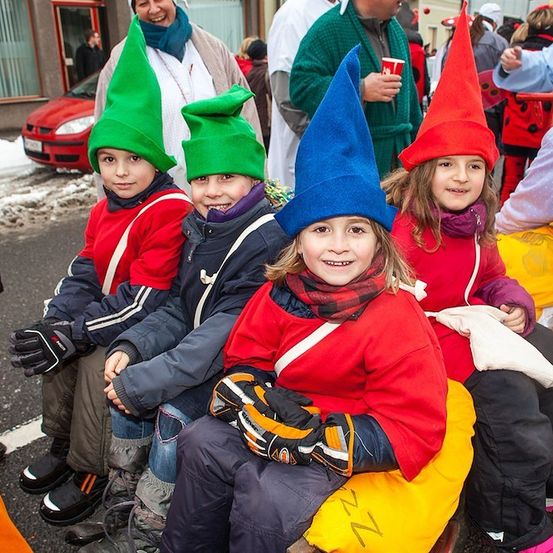Vier Kinder in passenden roten und blauen Kostümen und spitzen Hüten sitzen zusammen auf einer Bank in einer verschneiten Straße. Hinter ihnen stehen eine Frau in einem grünen Gewand und ein Mann in einem grünen Gewand.