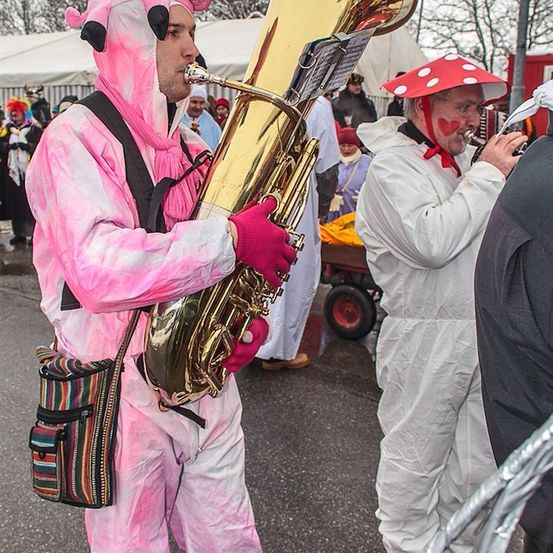 Ein Mann in einem rosa Schweinekostüm spielt Tuba, während eine Person in einem Pilzhut in der Nähe steht.