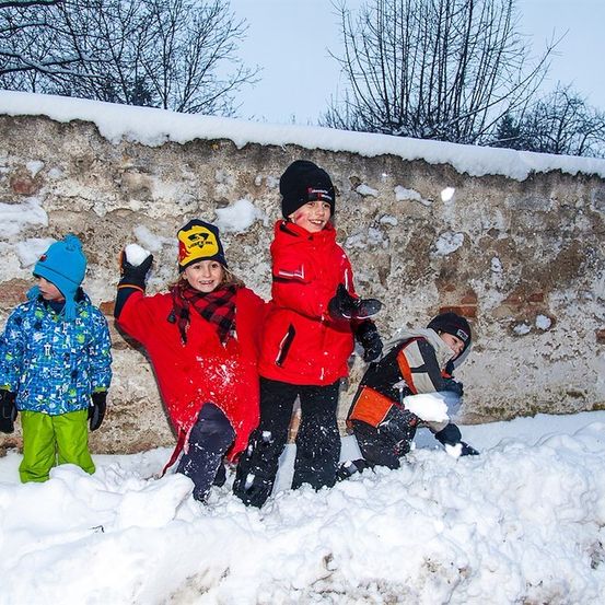 Vier Kinder in Winterkleidung spielen im Schnee vor einer Steinmauer.