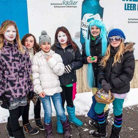 Eine Gruppe junger Mädchen in Winterkleidung und mit bemalter Gesichtsfarbe steht vor einer blau-weißen Wand mit Werbung.