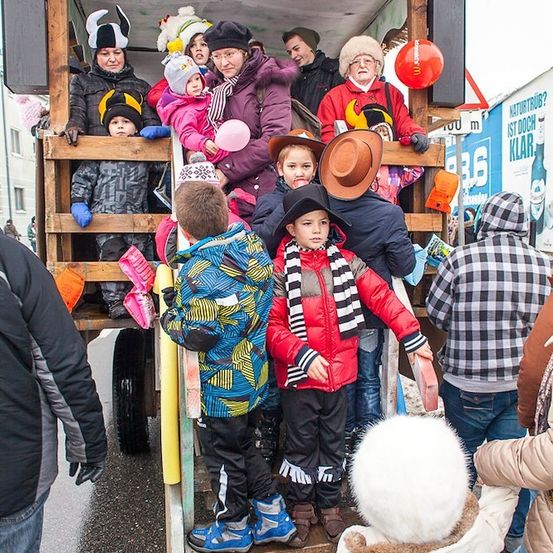 Eine Gruppe von Menschen, darunter Kinder, steht auf einem dekorierten Lastwagen, einige tragen Hüte und Schals, während andere Luftballons halten. Der Lastwagen ist von Menschen umgeben, und ein Schild ist im Hintergrund sichtbar.