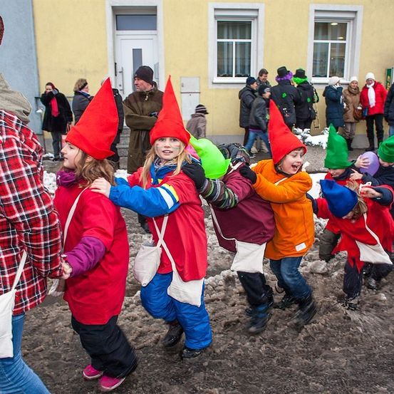 Eine Gruppe von Kindern in roten und blauen Kostümen mit spitzen Hüten geht in einer Reihe, möglicherweise für einen Umzug. Sie halten sich an den Händen. Dahinter stehen Menschen und beobachten.