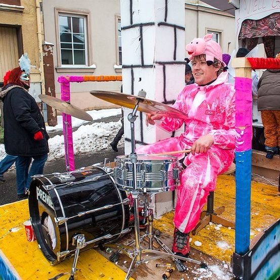 Ein Mann in einem rosa Schweinekostüm spielt die Trommeln auf einem Festwagen. Hinter ihm geht eine Frau mit roter Perücke und Maske an einem Gebäude vorbei.