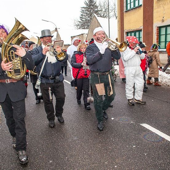 Eine Gruppe von Musikern in Kostümen marschiert eine verschneite Straße entlang und spielt Blechblasinstrumente.
