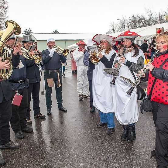 Eine Gruppe von Musikern in bunten Kostümen spielt Instrumente im Freien. Einige tragen Hüte und Stiefel. Der Hintergrund zeigt eine verschwommene Ansicht eines Gebäudes und Bäume.