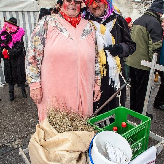 Zwei Personen in Kostümen und Schminke stehen neben einem Schubkarren mit Heu und einem Eimer. Dahinter sind weitere Personen ebenfalls in Kostümen gekleidet.