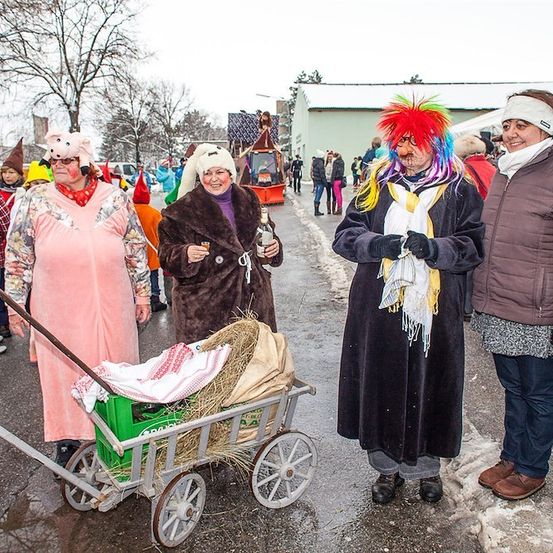 Eine Gruppe von Menschen in Kostümen und Mützen steht draußen im Schnee. Eine Frau schiebt einen Wagen mit Heu und einer Babypuppe. Eine andere Frau hält eine Flasche und einen Becher.