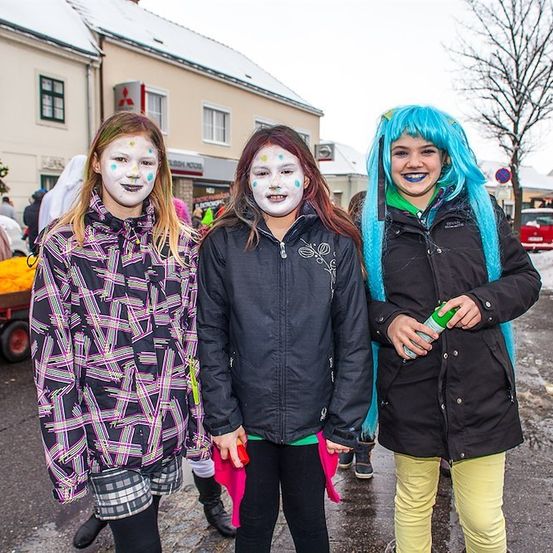 Drei Mädchen in Kostümen stehen auf einer verschneiten Straße. Das Mädchen auf der linken Seite hat bunte Gesichtsbemalung, die mittlere hat weiße Gesichtsbemalung und das Mädchen auf der rechten Seite hat blaue Haare und eine grüne Flasche. Dahinter befinden sich Gebäude, ein Baum und ein Wagen mit gelben Objekten.