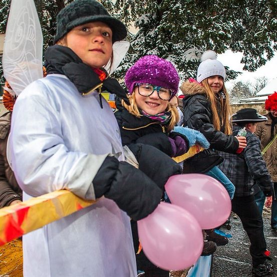 Eine Gruppe von Kindern in Winterkleidung und mit rosa Luftballons in der Hand geht eine verschneite Straße entlang. Ein Kind trägt ein weißes Feenkostüm mit Flügeln.
