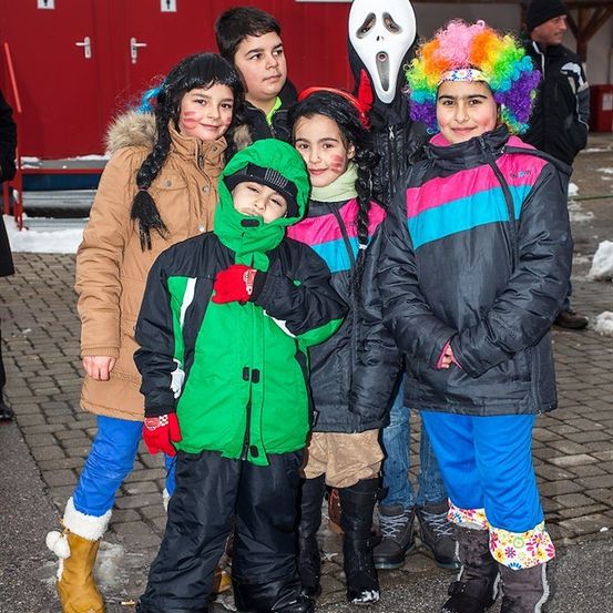 Eine Gruppe von Kindern in farbenfrohen Wintermänteln und Stiefeln, zwei von ihnen mit Gesichtsbemalung, stehen zusammen in einer verschneiten Außenumgebung.