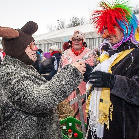 Zwei Personen in farbenfrohen Kostümen, eine als Maus und die andere mit Clownsmaske, schütteln sich die Hände im Freien. Hinter ihnen stehen mehrere Personen, einige mit Tiermasken. Im Vordergrund ist ein grüner Kasten mit Flaschen zu sehen.