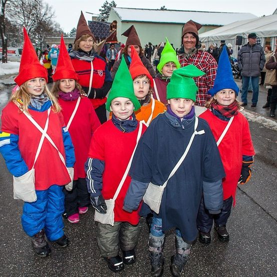 Eine Gruppe von Kindern in roten und blauen Kostümen mit kegelförmigen Hüten steht auf einer Straße. Hinter ihnen sind Erwachsene in verschiedenen Outfits anwesend, und einige beobachten sie. Der Hintergrund zeigt einen verschneiten Bereich mit Bäumen und einem Gebäude.