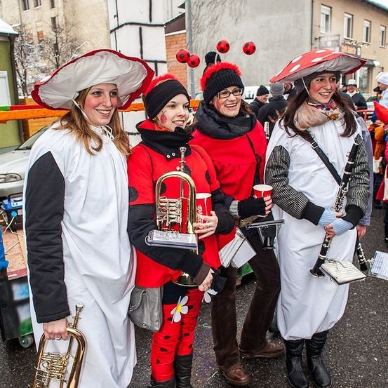 Vier Frauen in Kostümen, zwei mit Trompeten, eine mit Klarinette und eine mit Flöte, stehen auf einer Straße mit Gebäuden im Hintergrund.