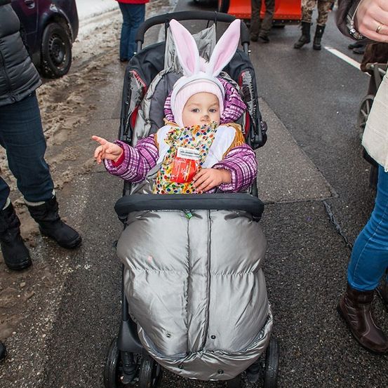 Ein Baby in einem Hasenkostüm sitzt in einem Kinderwagen auf einer verschneiten Straße, umgeben von Menschen und einem Auto.