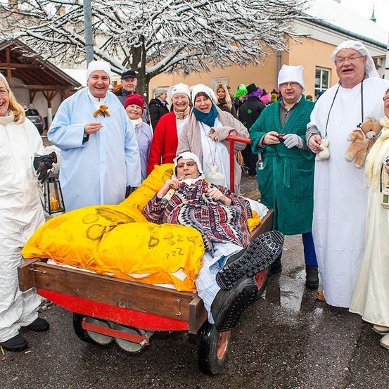 Eine Gruppe von Menschen in verschiedenen Kostümen, darunter ein Krankenhauskittel, versammelt sich um eine Person in einem roten Wagen. Sie lächeln und scheinen an einem festlichen Ereignis teilzunehmen.