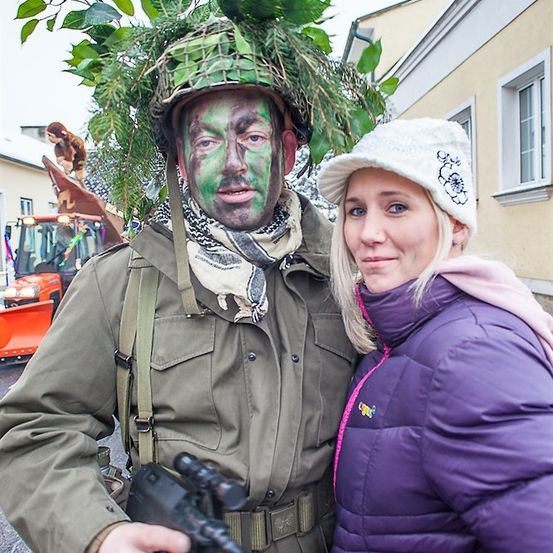 Ein Mann, verkleidet als Soldat mit einem Tarnhelm und Gesichtsbemalung, steht neben einer Frau in einer lila Jacke.