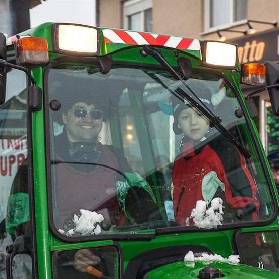Ein Mann und ein Kind sitzen in einem grünen Traktor, lächeln und haben Schnee auf der Windschutzscheibe. Sie befinden sich in einer verschneiten Umgebung im Freien.