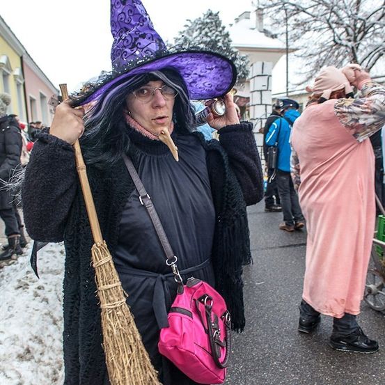 Eine Frau in einer Hexenverkleidung mit einem lila Hut und einer Besen, die auf einer verschneiten Straße steht, mit Leuten um sie herum.