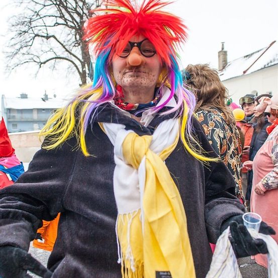 Ein Mann mit mehrfarbigen Haaren, der einen gelben Schal und einen schwarzen Mantel trägt und einen Becher in der Hand hält, bei einer festlichen Veranstaltung im Freien.