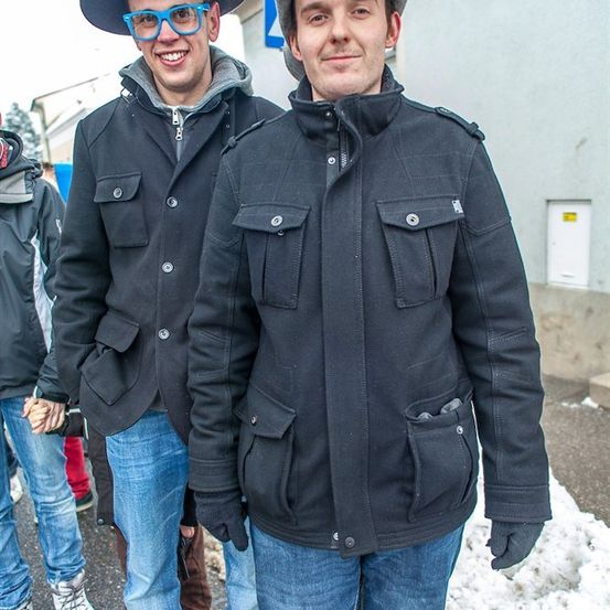 Zwei Männer stehen nebeneinander und tragen beide schwarze Jacken und Hüte. Sie lächeln und scheinen für ein Foto zu posieren. Der Mann auf der linken Seite trägt eine Brille und Handschuhe.
