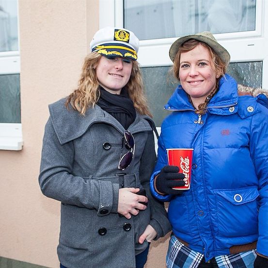 Zwei Frauen stehen vor einem Gebäude, eine trägt eine dunkelblaue Pufferjacke und hält einen roten Coca-Cola-Becher, die andere ein graues Kleid mit einer dunkelblauen Mütze und Sonnenbrille an der Vorderseite.