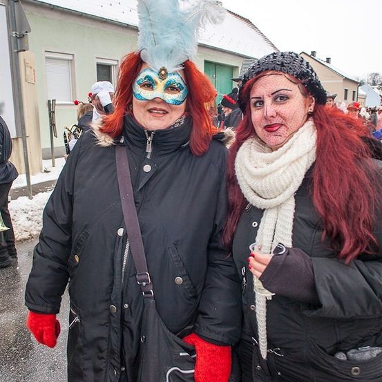 Zwei Frauen in Kostümen und Make-up, eine mit einer blauen Federmaske, stehen auf einer verschneiten Straße.