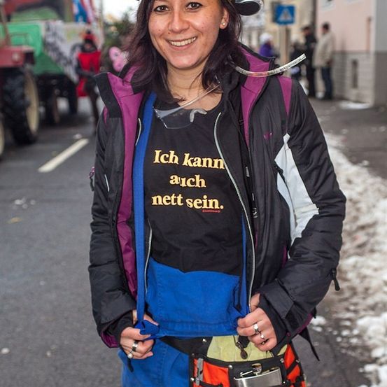 Eine Frau mit einer gelben Dose auf dem Kopf lächelt auf der Straße. Sie trägt ein schwarzes T-Shirt mit dem Text 'Ich kann auch nett sein'. Hinter ihr stehen ein Traktor und einige Leute am Straßenrand.