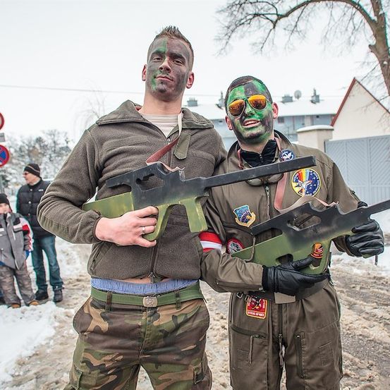 Zwei Männer in Camouflage-Uniformen und Gesichtsbemalung stehen im Schnee und halten eine große Spielzeugpistole. Dahinter stehen einige Leute auf dem verschneiten Boden.