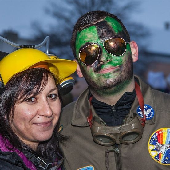 Eine Frau mit einem gelben Helm und ein Mann mit grüner Gesichtsbemalung und Schutzbrille stehen zusammen.